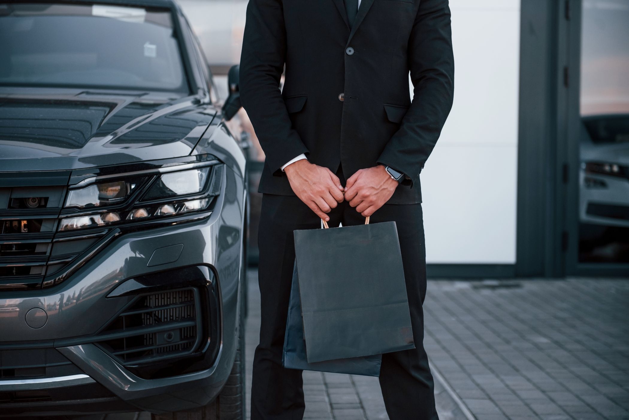 A well dressed man in a black suit with high end shopping bags in his hands stands before a luxury car in Miami.
