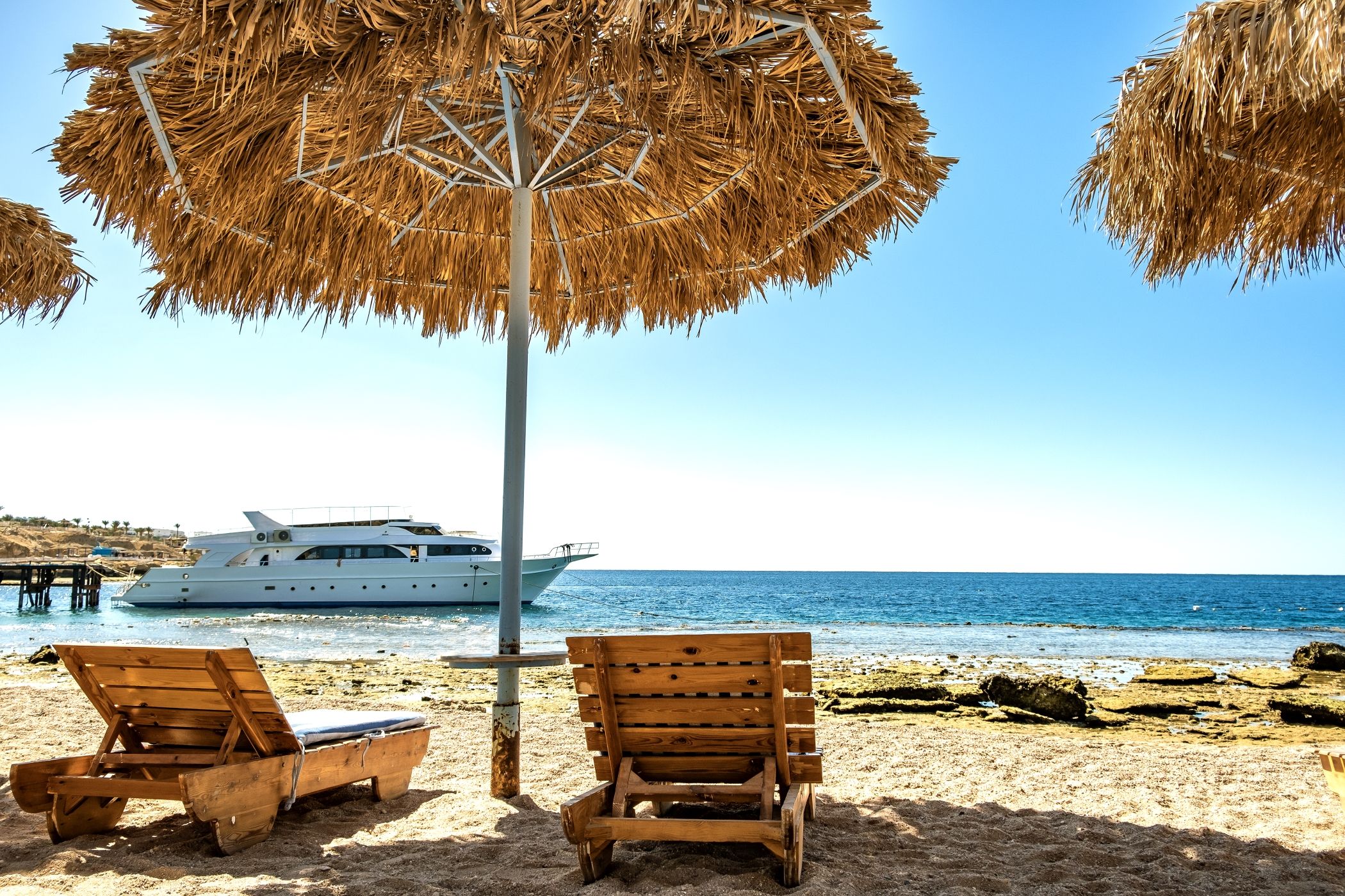 Wooden deck chairs under rough straw sun umbrella on sea beach and big white yacht ship in water near Miami.