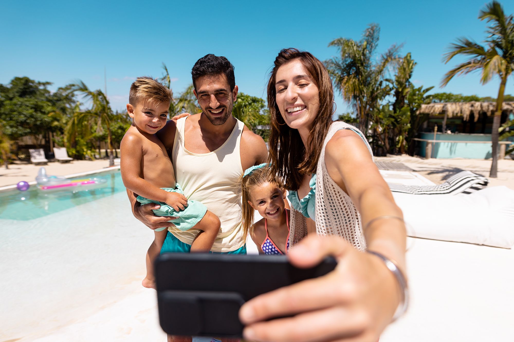A smiling family of four taking a selfie with palm trees and a swimming pool in the background.