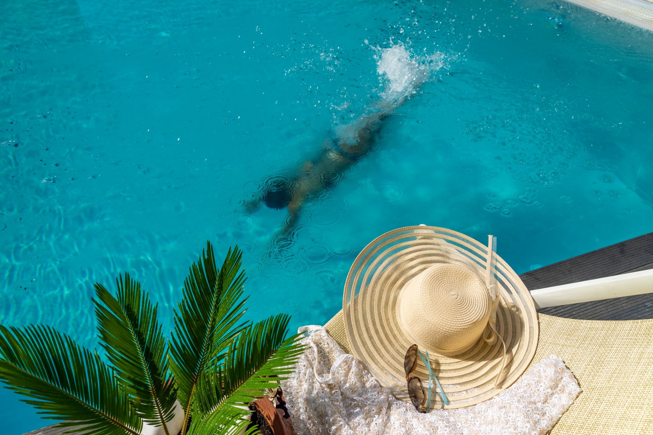 Woman swimming underwater in a luxury villa in Miami, a hat, sunglasses and plan are on the edge of the pool.